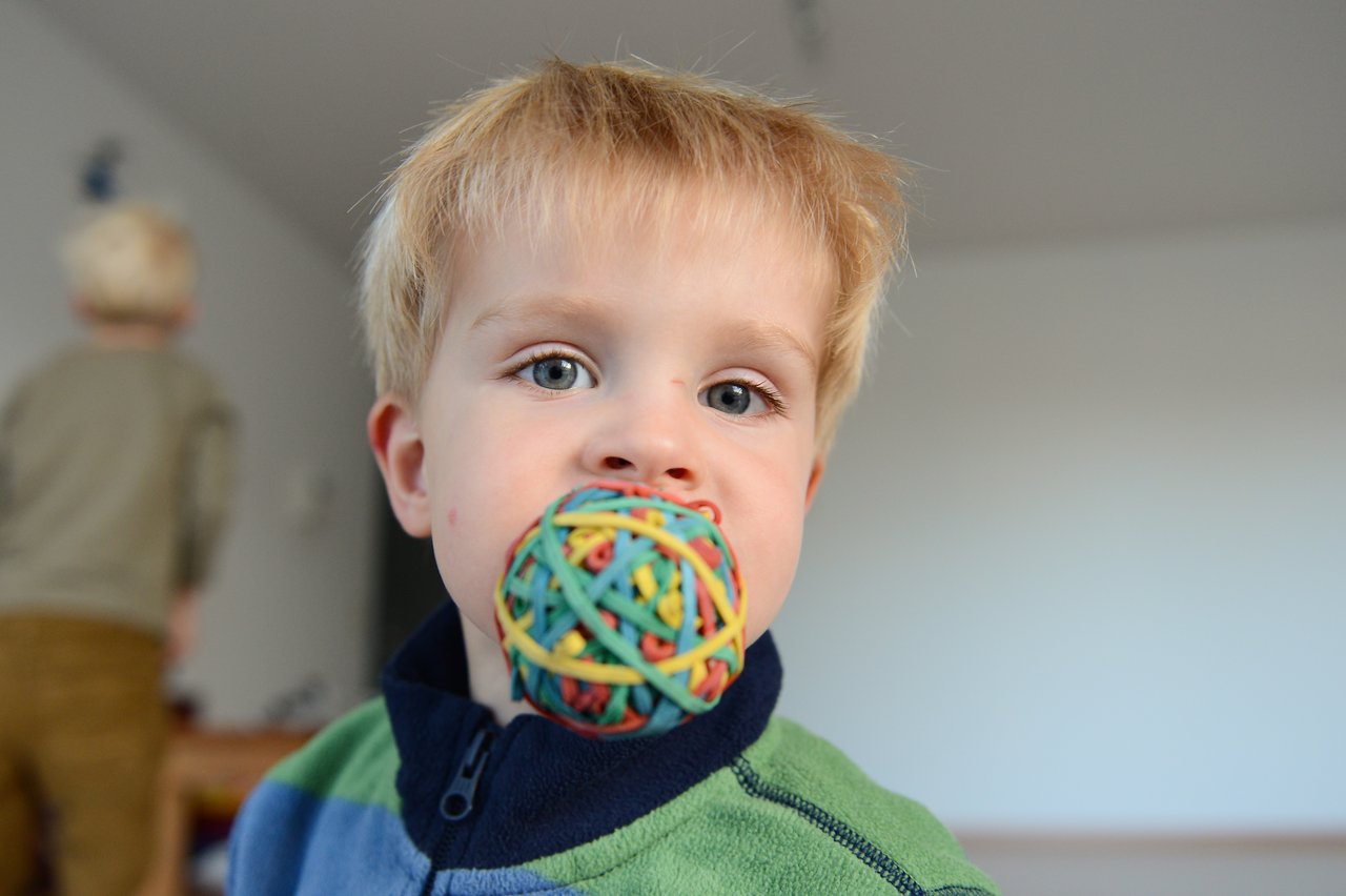A young child holds a colorful rubber band ball in their mouth, looking directly at the camera.