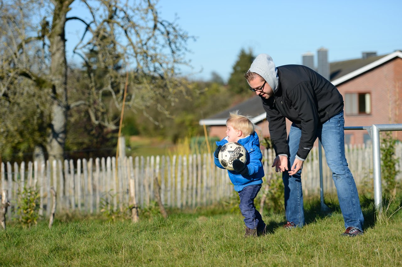 A man in a hoodie helps a young child holding a soccer ball in a grassy outdoor area.