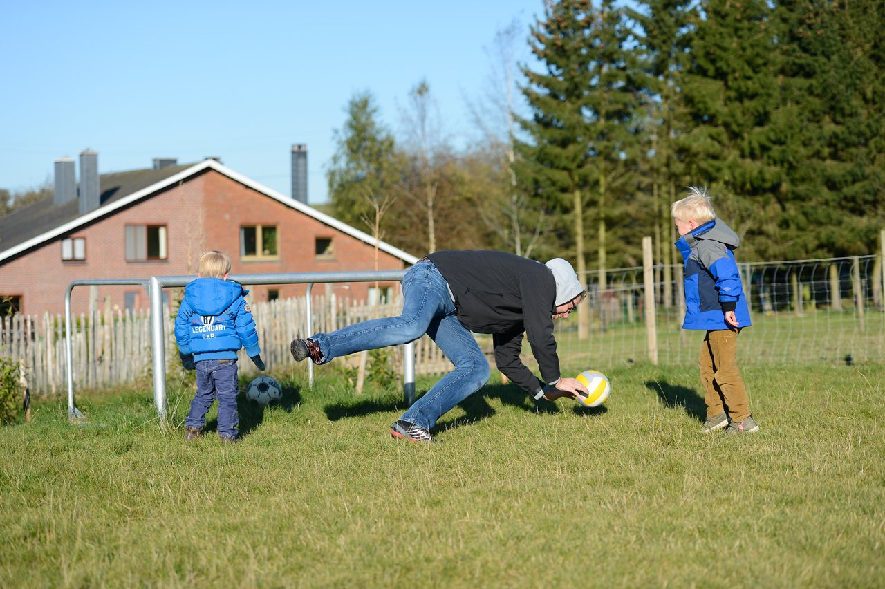 An adult in a hoodie bends down to catch a volleyball while two children watch on a grassy field.
