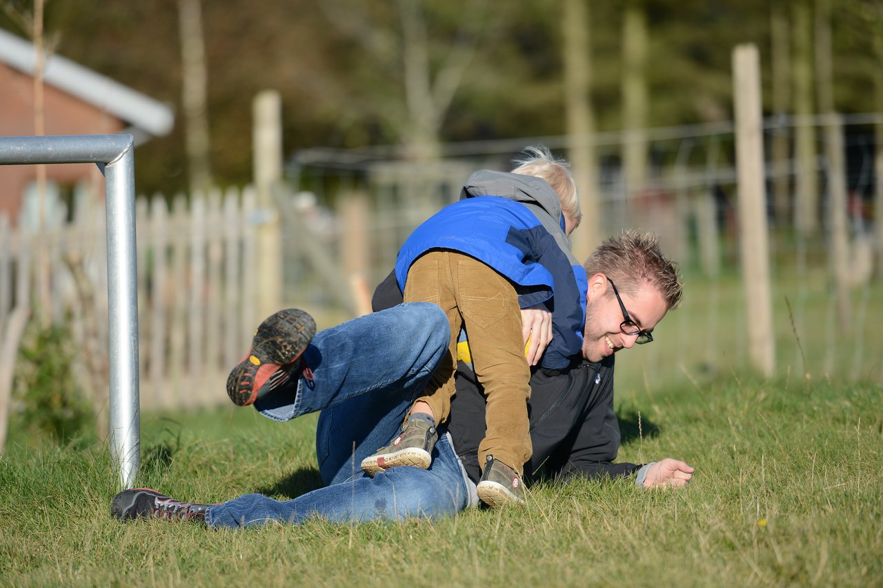 A child in a blue jacket climbs on top of a smiling man lying on the grass.