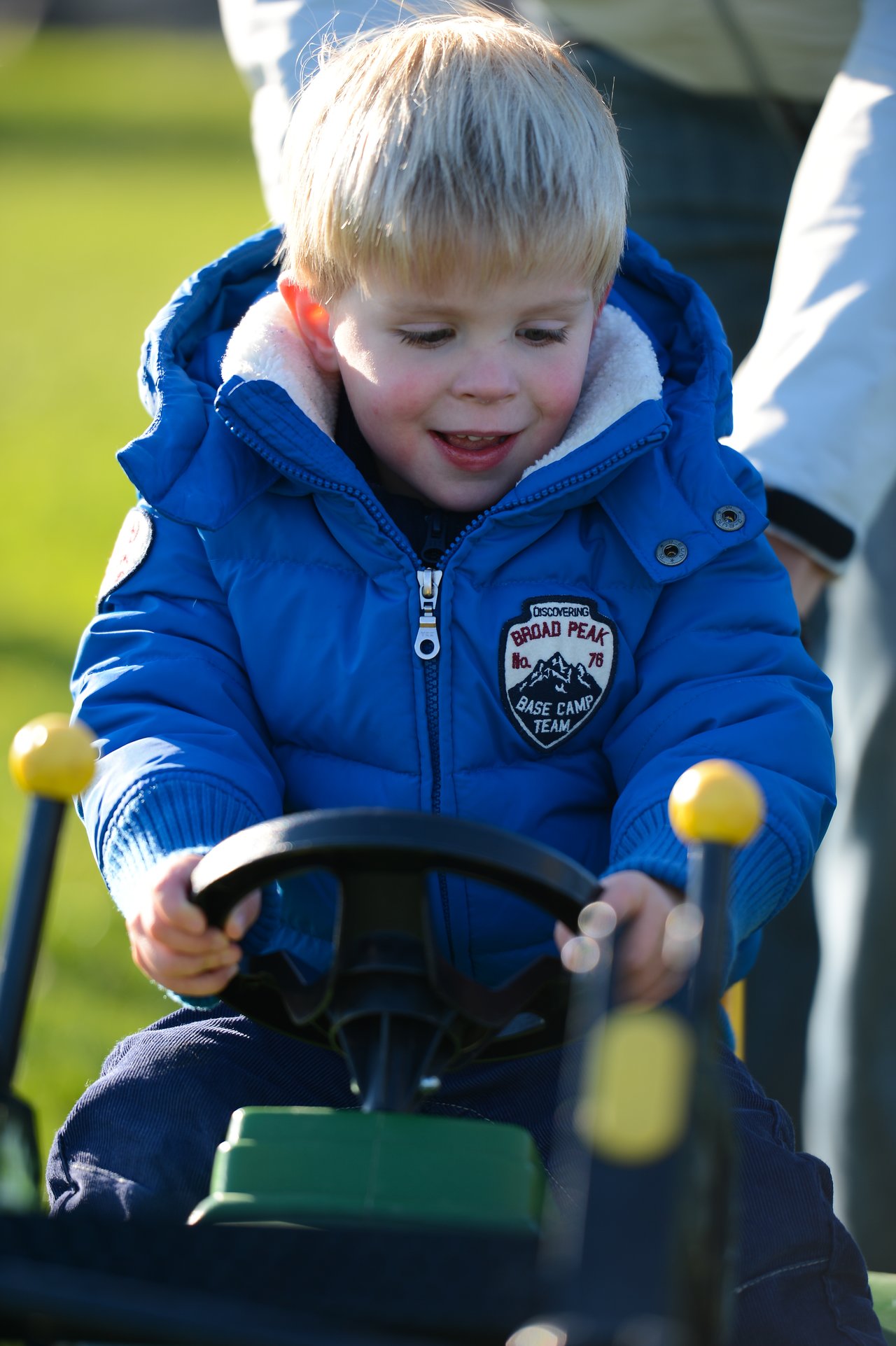 A young child in a blue jacket grips the steering wheel of a small toy vehicle while an adult assists.