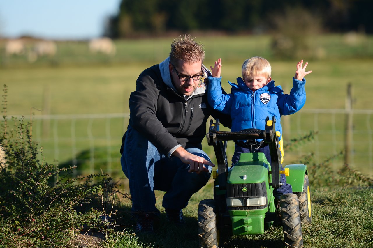 A man crouches beside a child on a toy tractor, pointing while the child raises their hands.