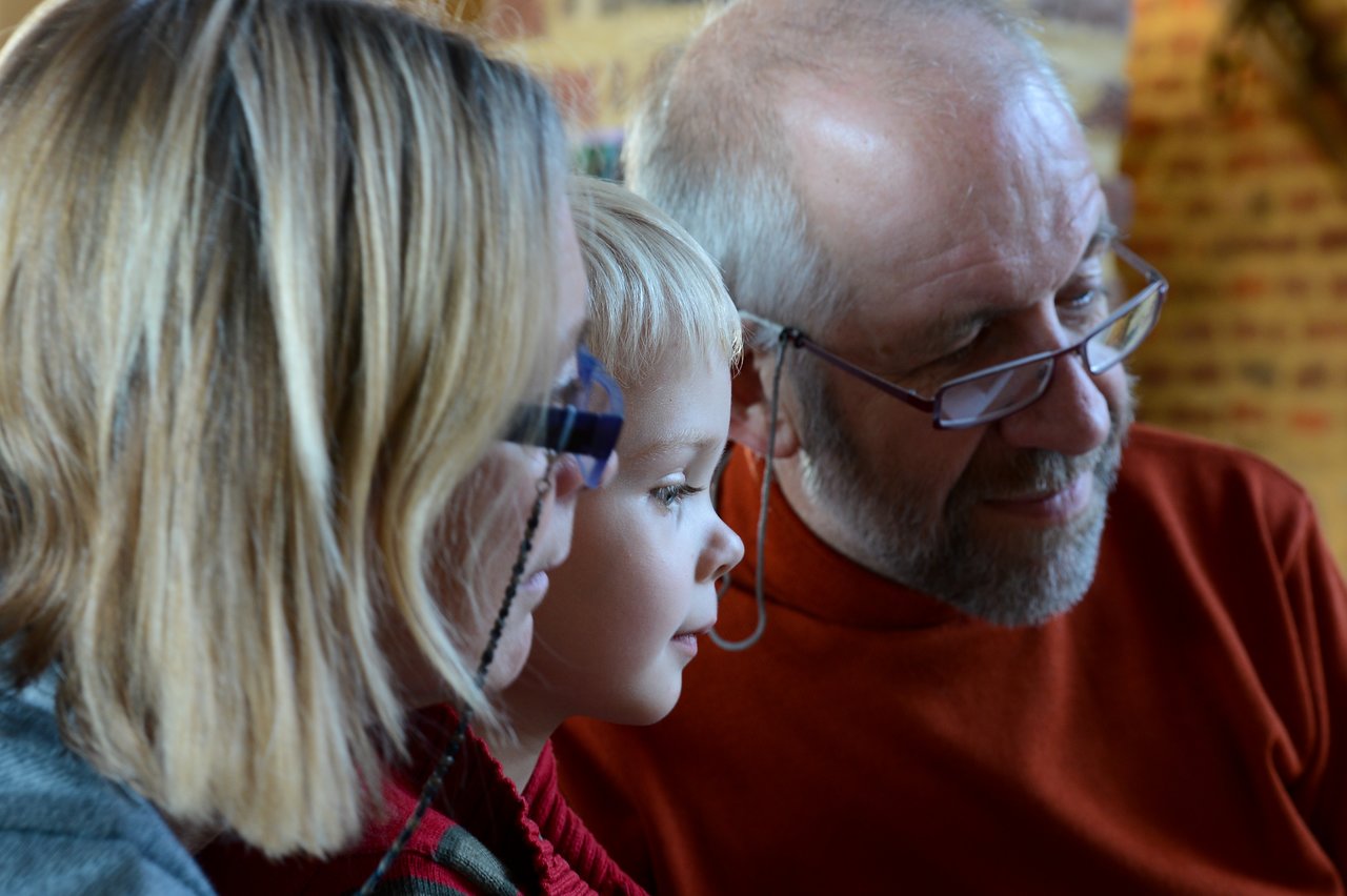 A child sits between two adults, all looking at something closely with focused expressions.