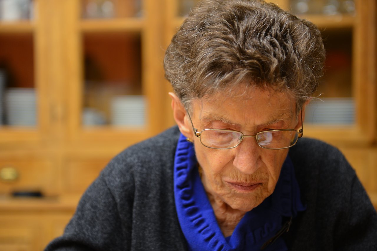 An elderly woman wearing glasses and a dark sweater looks down with focus, possibly reading or writing.
