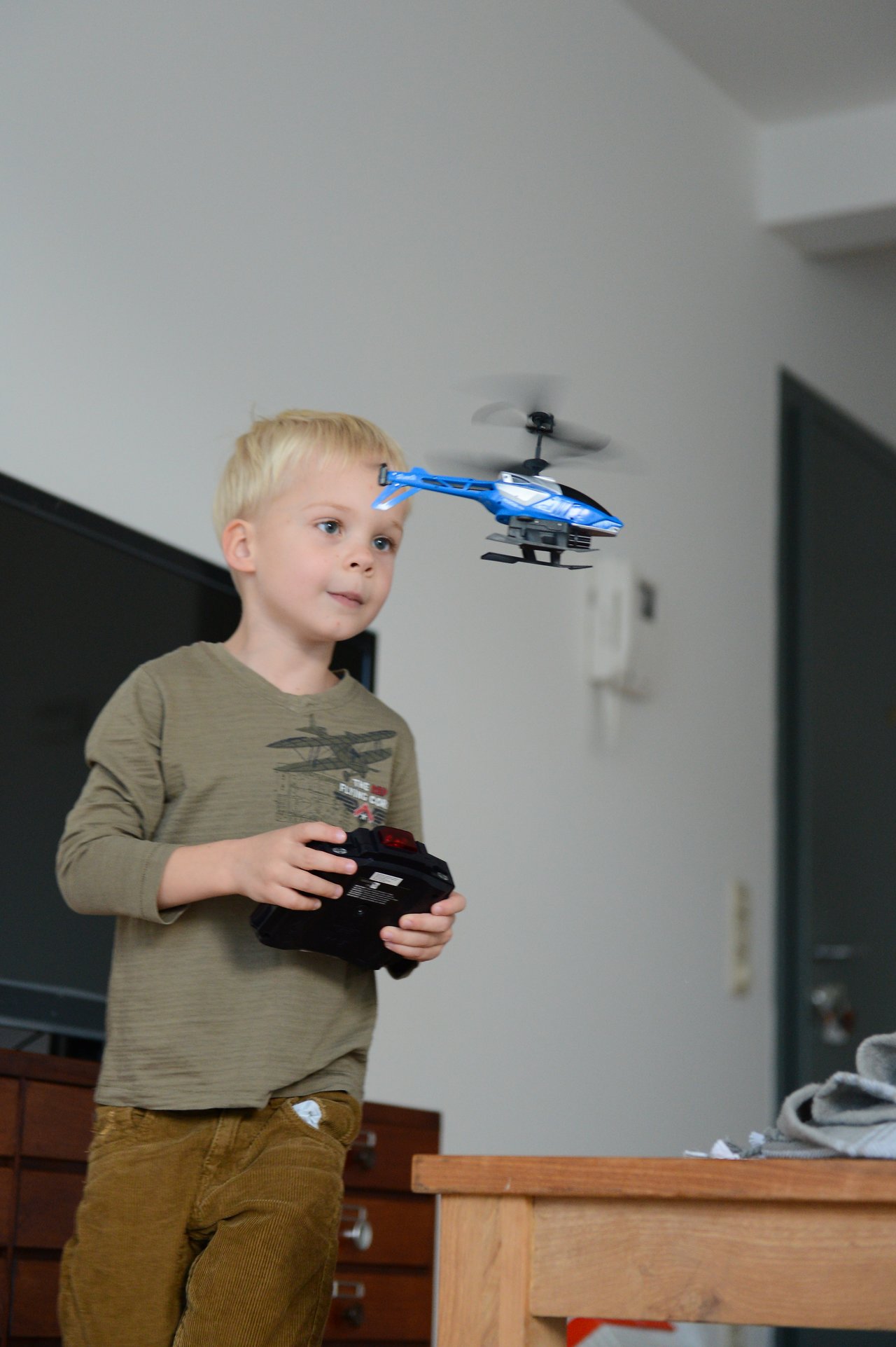 A young boy controls a flying toy helicopter with a remote, focusing intently indoors.