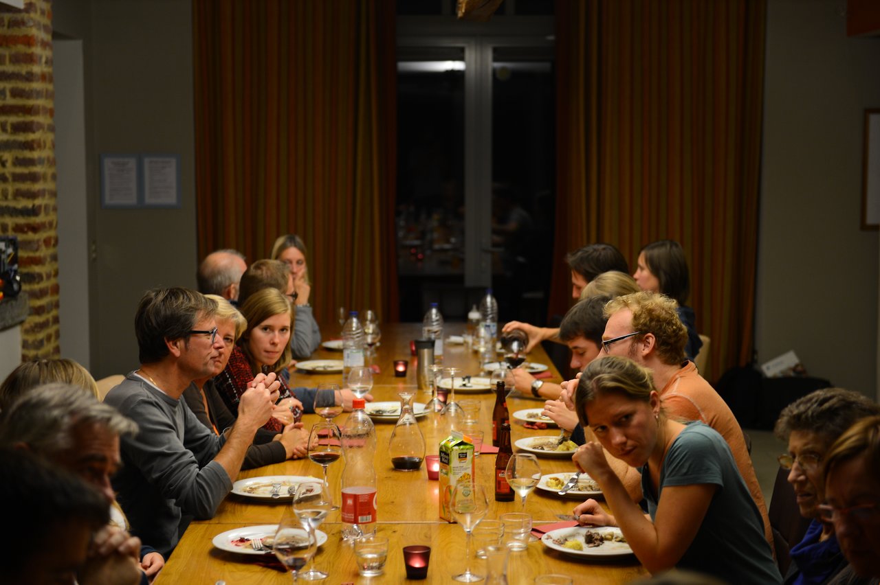 A group of people sit around a long table, eating, drinking, and talking during a meal.