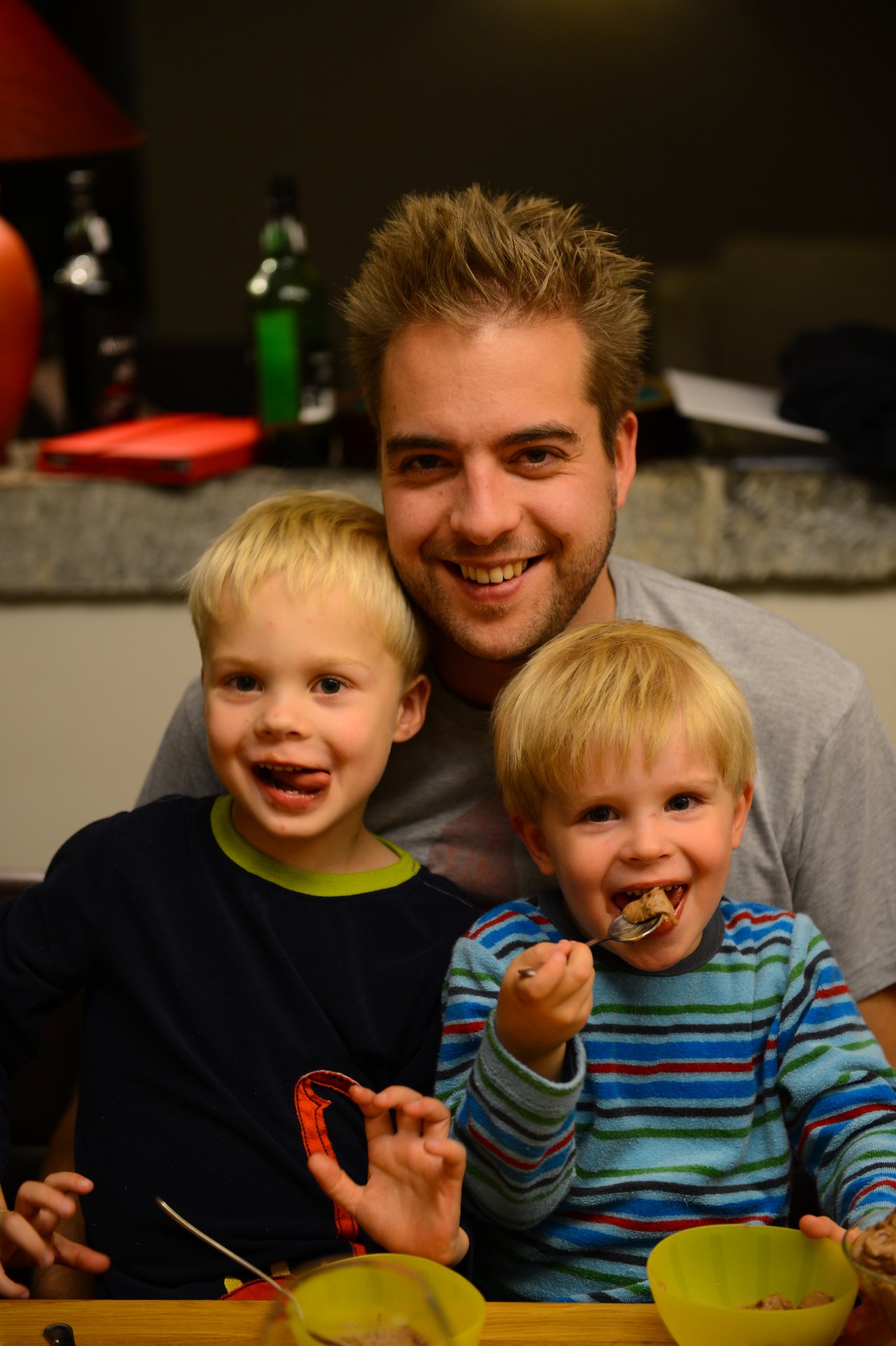 A smiling man sits behind two young children eating dessert, with one playfully sticking out their tongue.