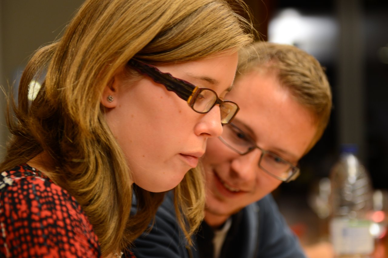 A woman wearing glasses focuses on something in front of her while a man beside her smiles and looks on.