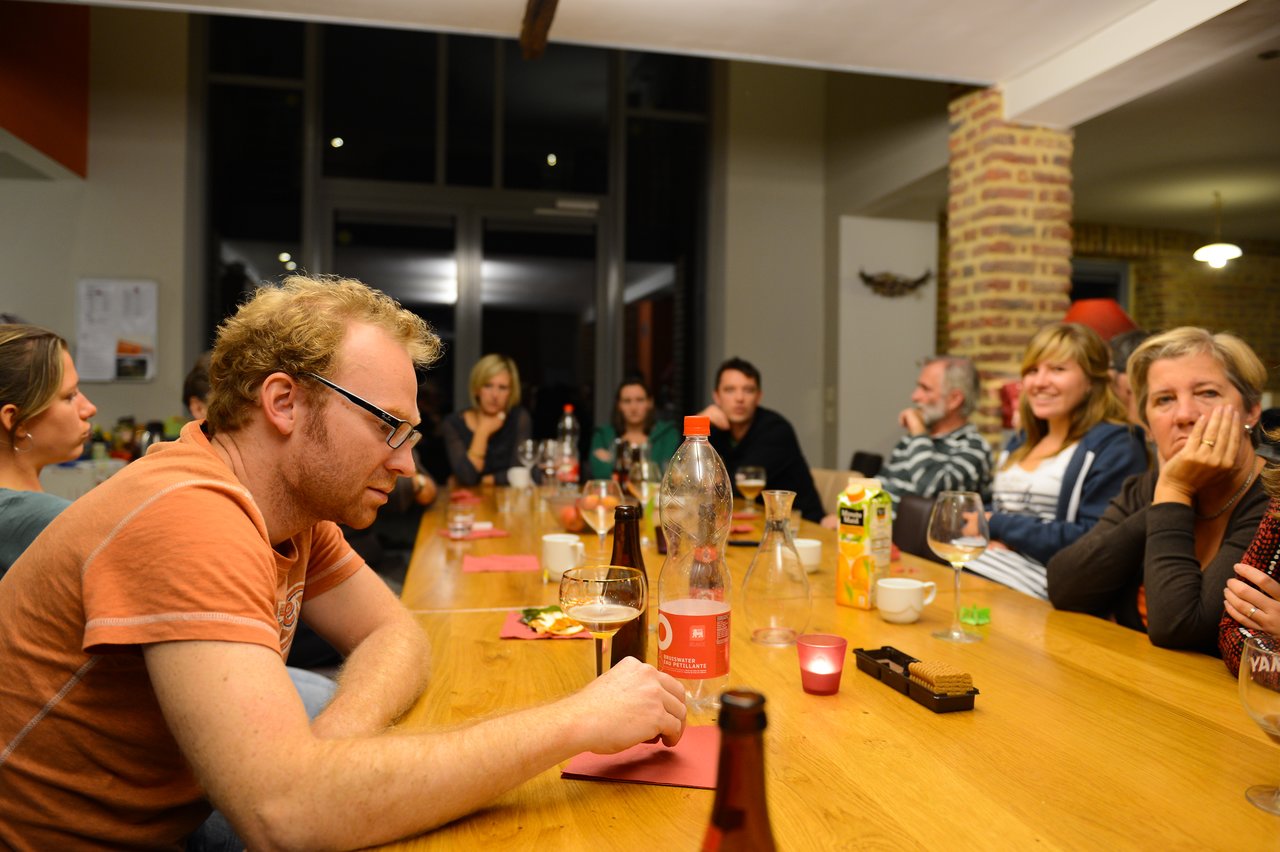 A man in glasses sits at a table with drinks and food, surrounded by a group of people socializing.