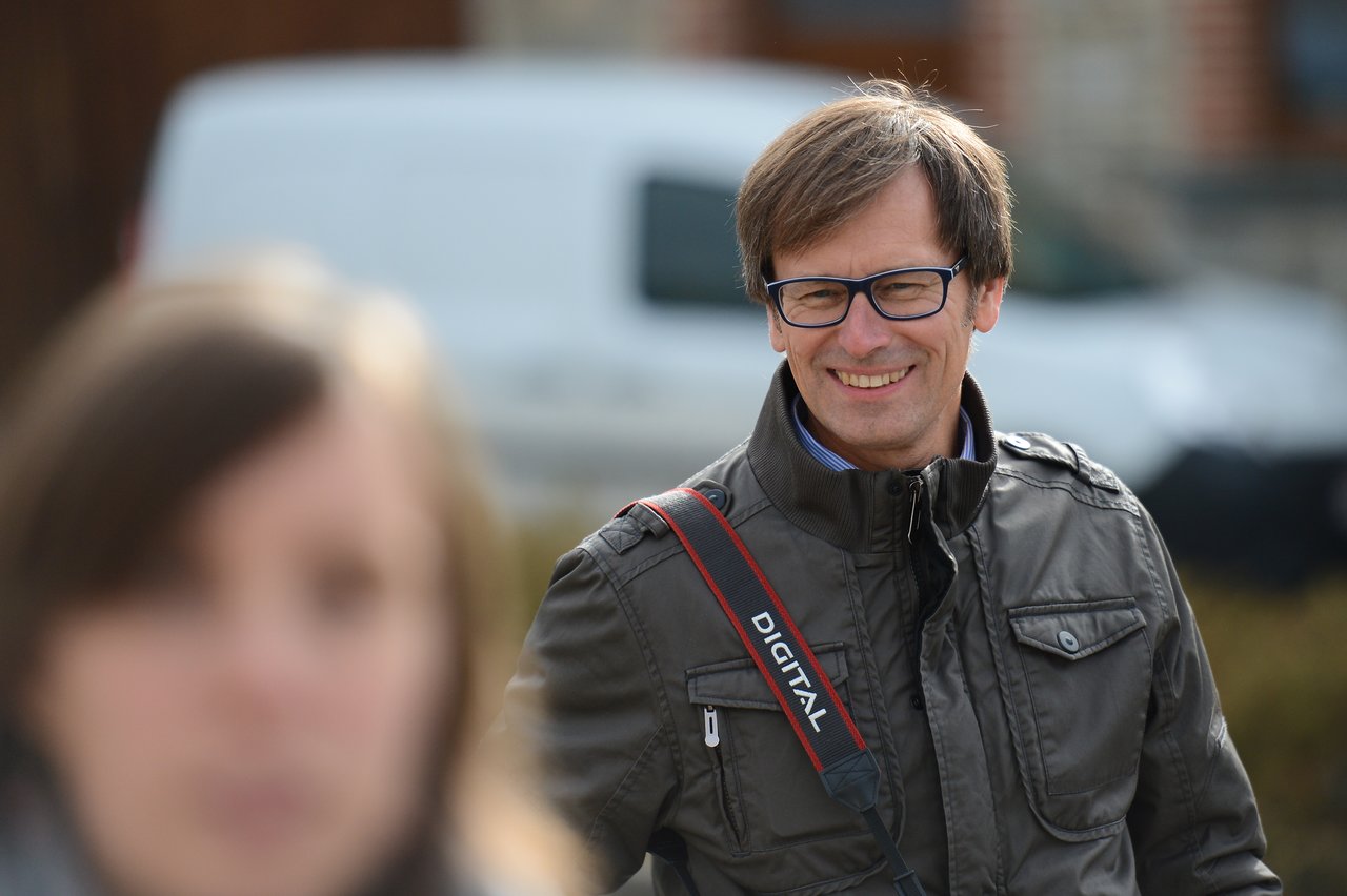 A man wearing glasses and a jacket smiles while carrying a camera strap.