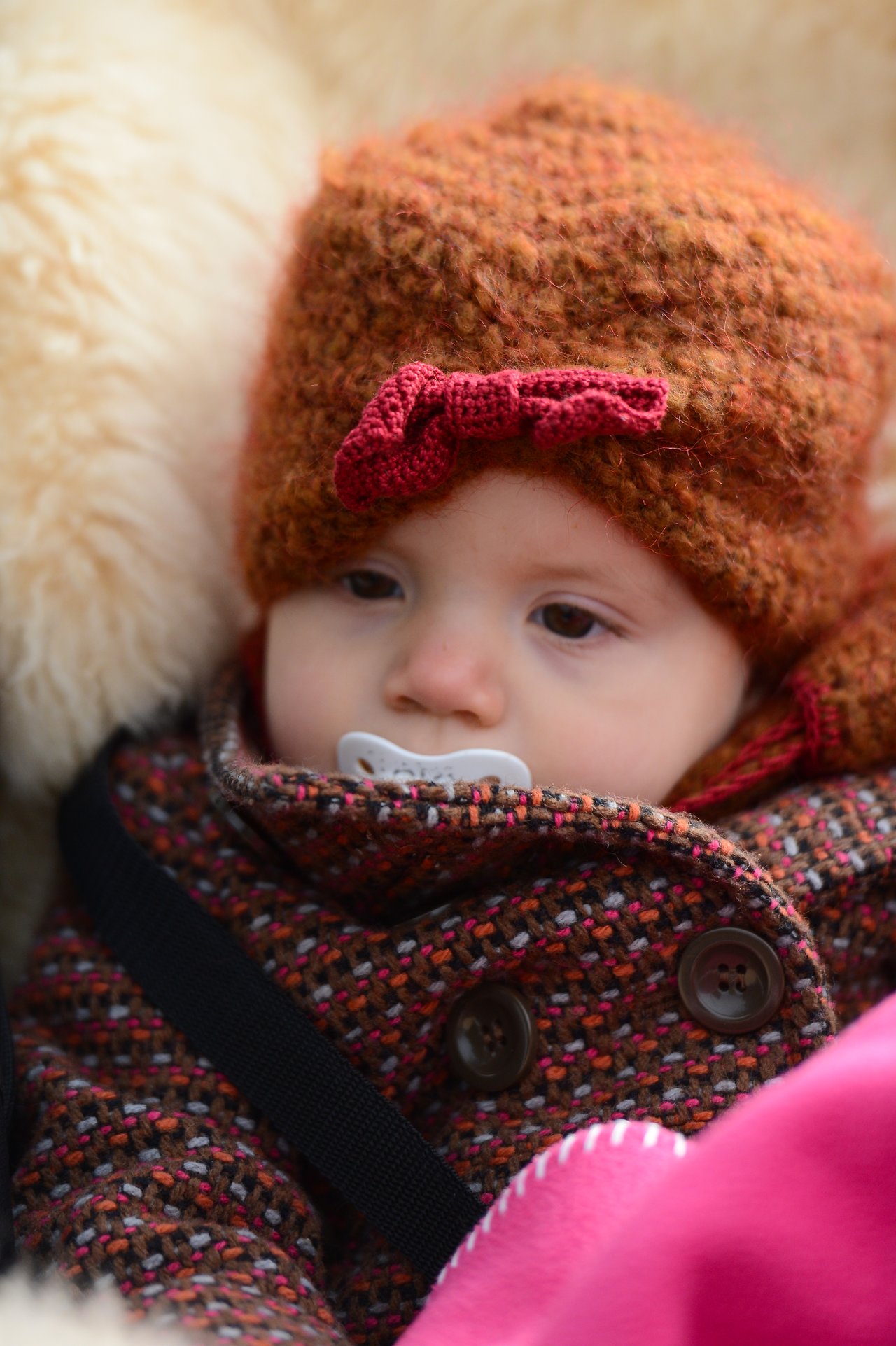 A baby wearing a warm hat and coat sits bundled up with a pacifier in their mouth.