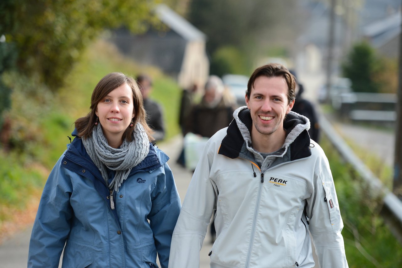 Two people in jackets and scarves walk outdoors, smiling at the camera, with others walking behind them.