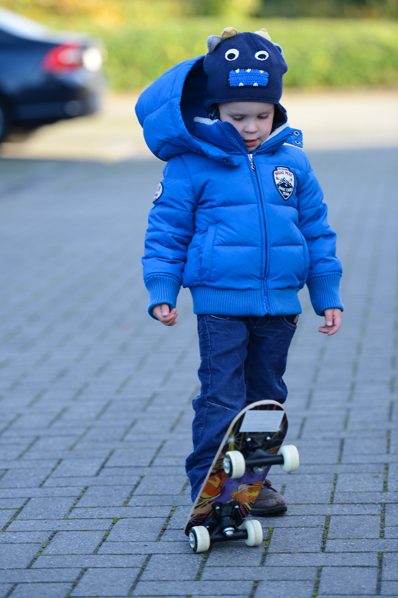 A child in a blue jacket and hat looks down at a skateboard tilted on its wheels.