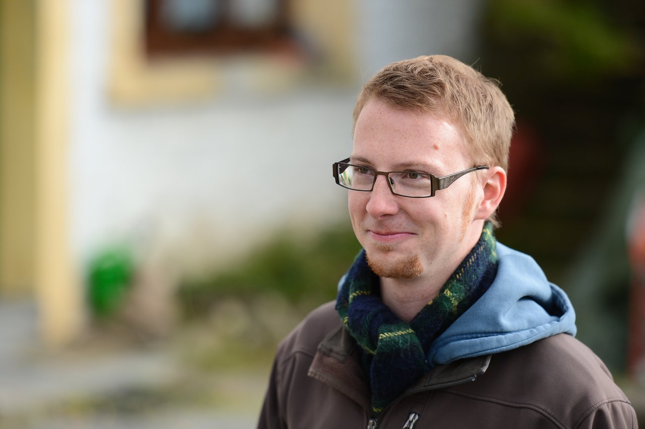 A man with glasses and a scarf smiles slightly while standing outdoors in a casual jacket.