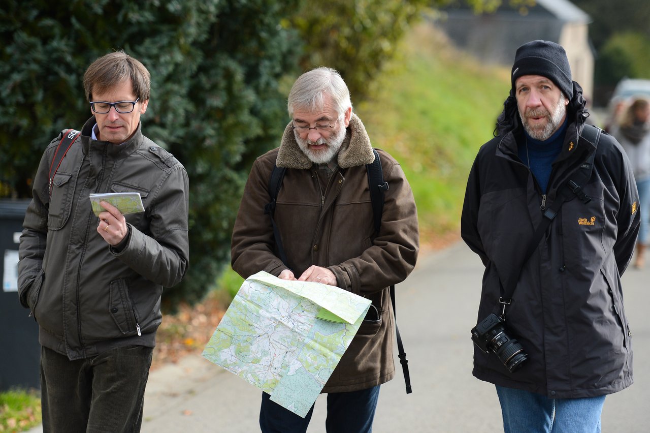 Three men walk along a path, two holding maps while the third carries a camera.