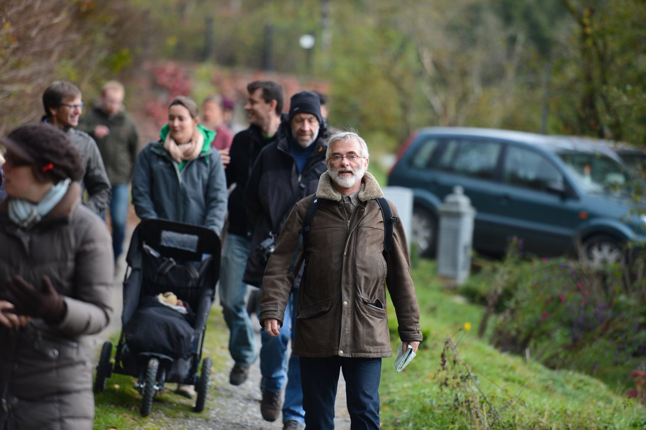 A group of people walks along a path, with a man in a brown jacket leading the way.