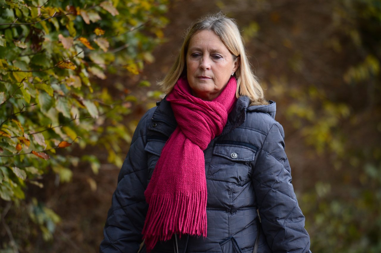 A woman in a dark coat and red scarf walks outdoors, looking down with a neutral expression.