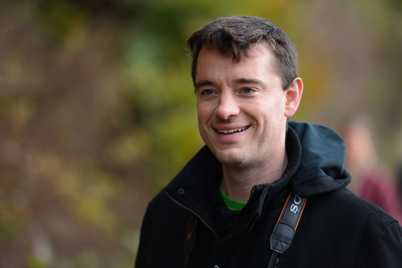 A man wearing a black jacket and a camera strap smiles while standing outdoors with a blurred background.