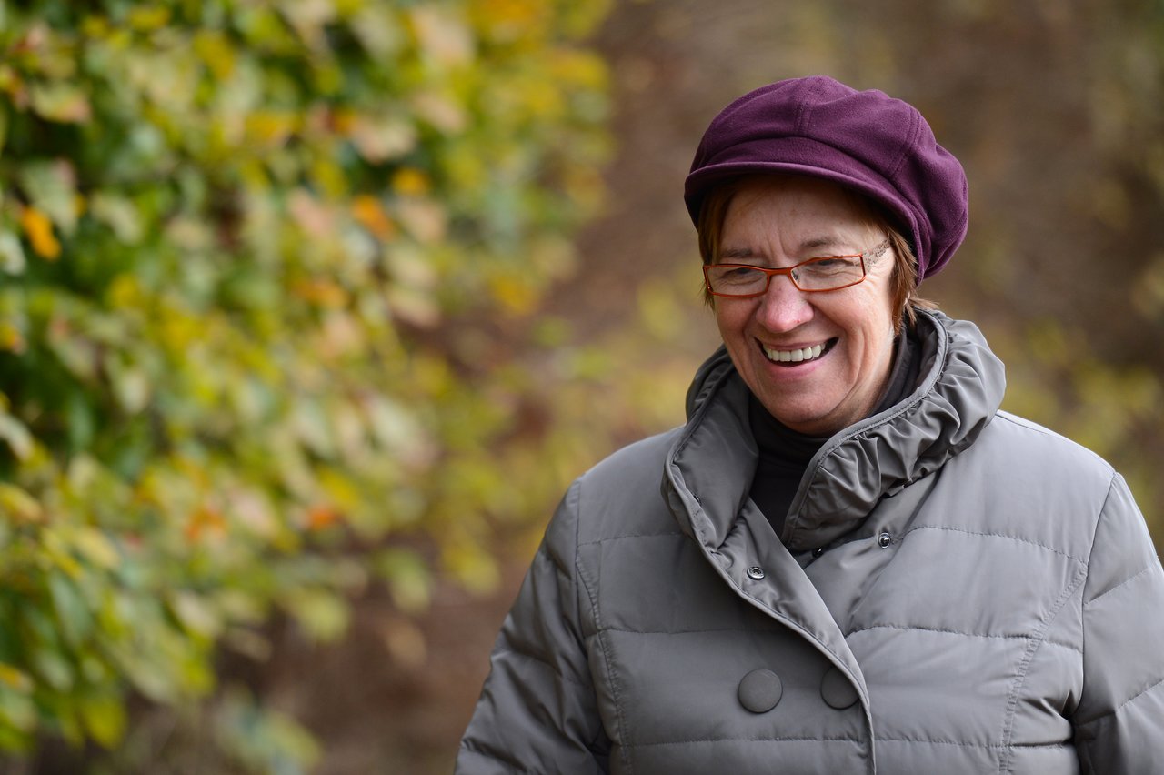 A woman wearing a purple hat and gray coat smiles while standing outdoors with blurred greenery in the background.