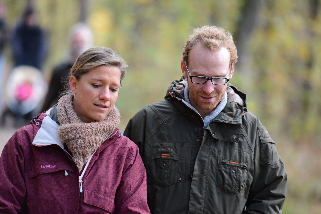 A man and a woman in warm jackets walk together outdoors, looking down and engaged in conversation.