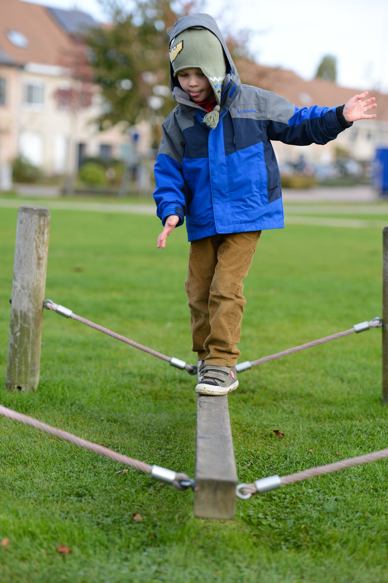 A child in a blue jacket balances on a wooden beam at a playground, arms outstretched for stability.