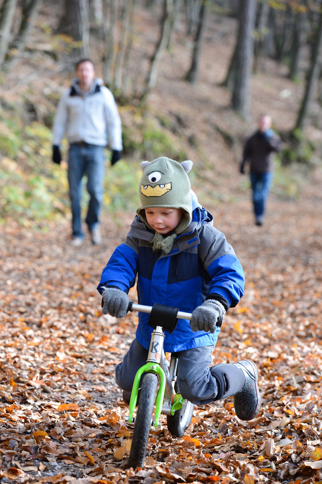 A child in a blue jacket rides a small balance bike on a leaf-covered path, with two adults behind.