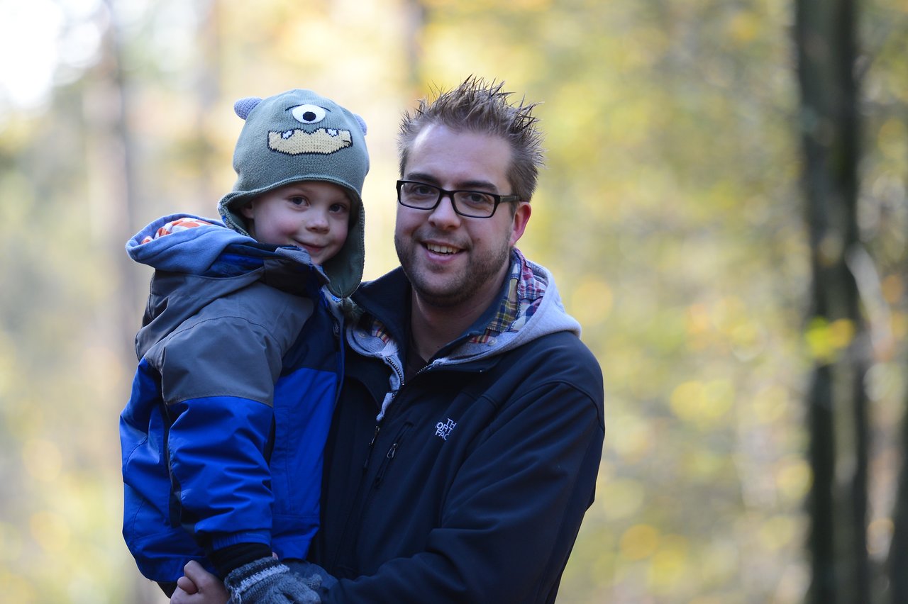 A man wearing glasses holds a young child in a blue jacket and monster hat outdoors.