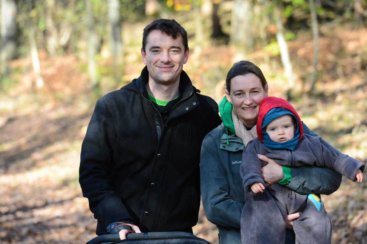 A smiling couple poses outdoors with their baby, who is dressed in a hooded outfit and held in their arms.
