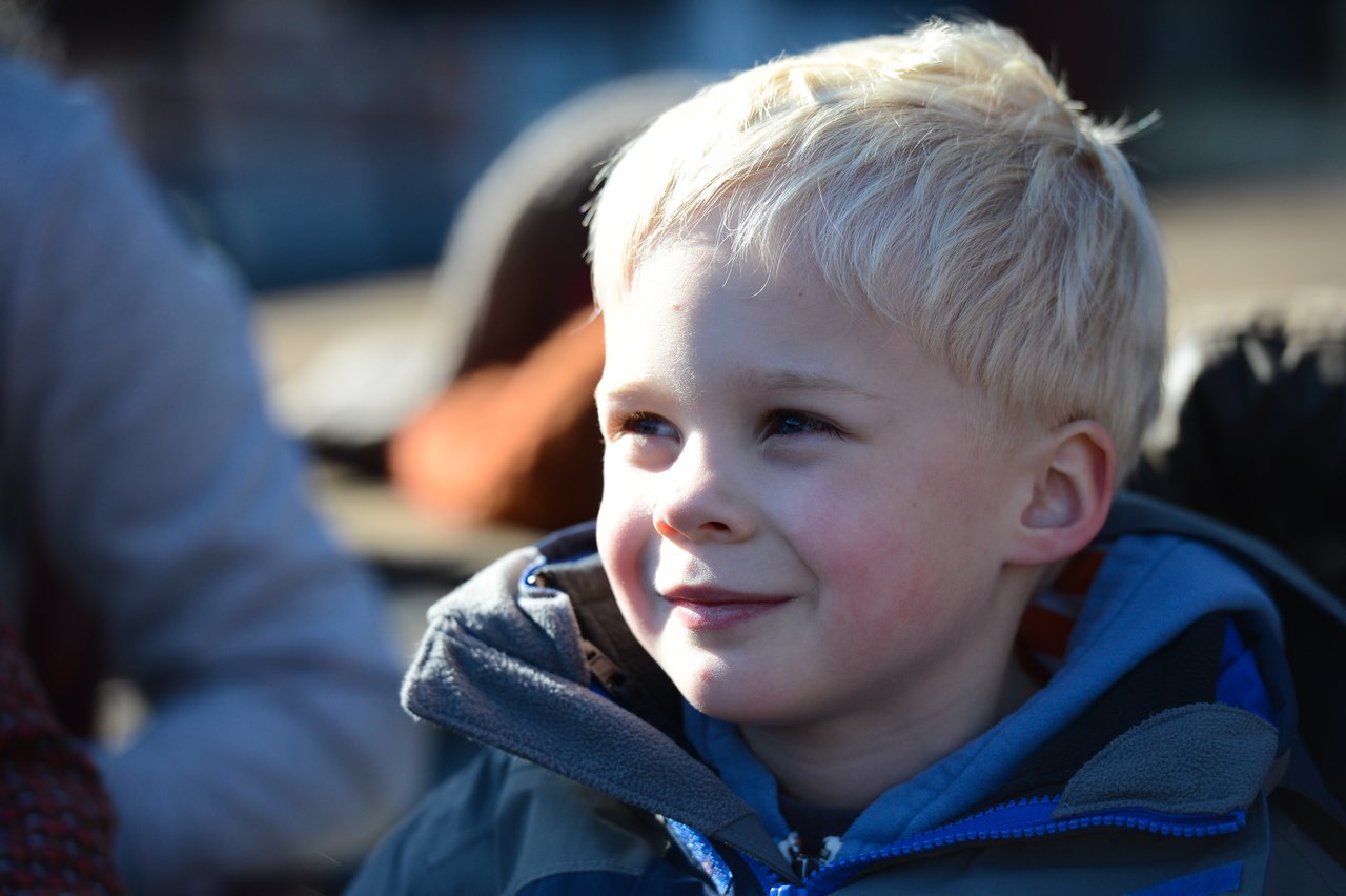 A young child with blond hair smiles while looking slightly to the side, wearing a blue jacket outdoors.
