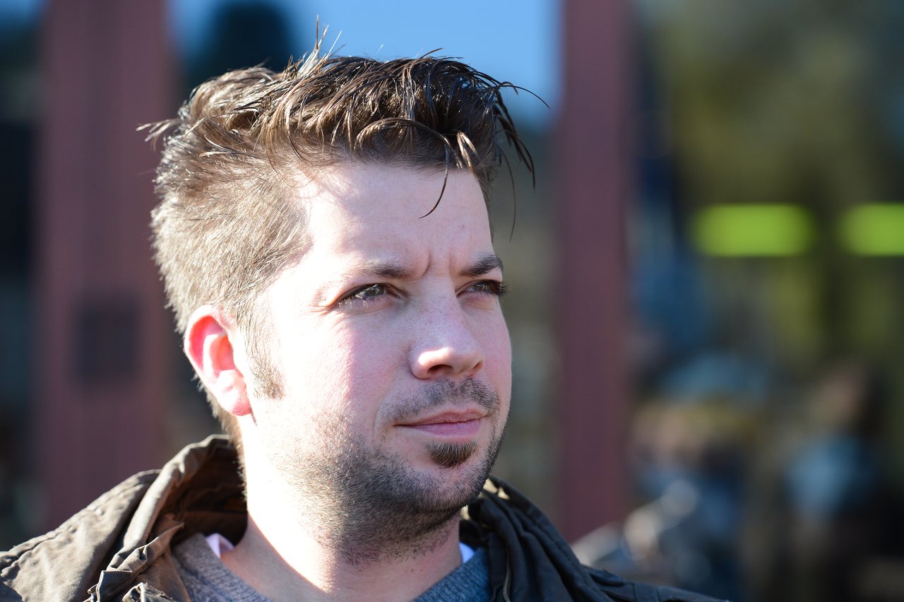 A man with short, styled hair looks off to the side in natural sunlight, wearing a jacket.