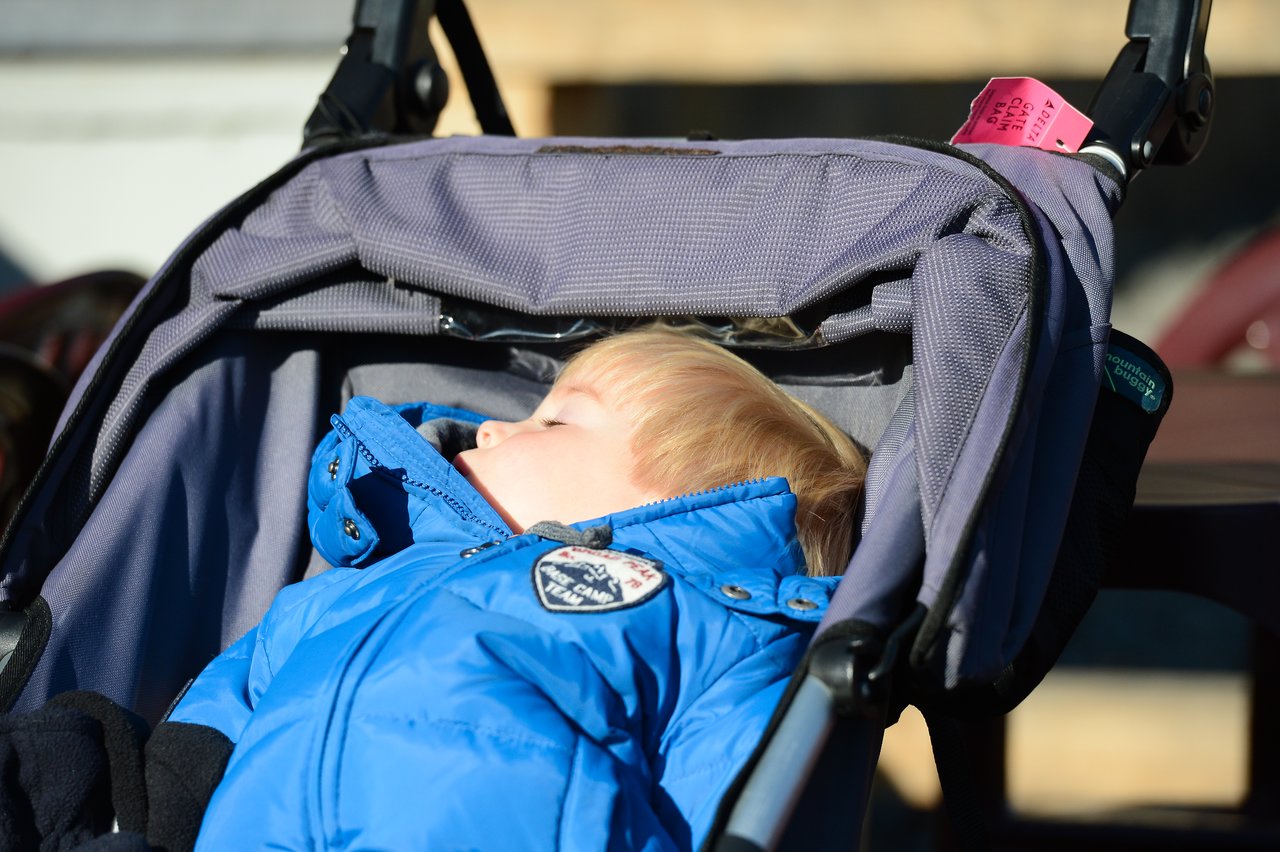 A young child in a blue jacket sleeps in a stroller under bright sunlight.