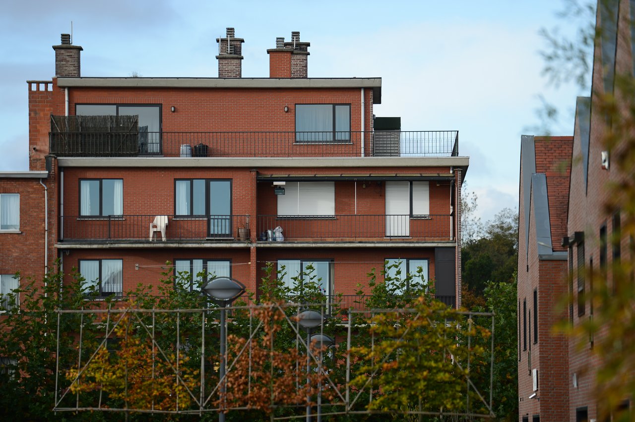 A red brick apartment building with multiple balconies, some with chairs and plants, surrounded by trees and streetlights.