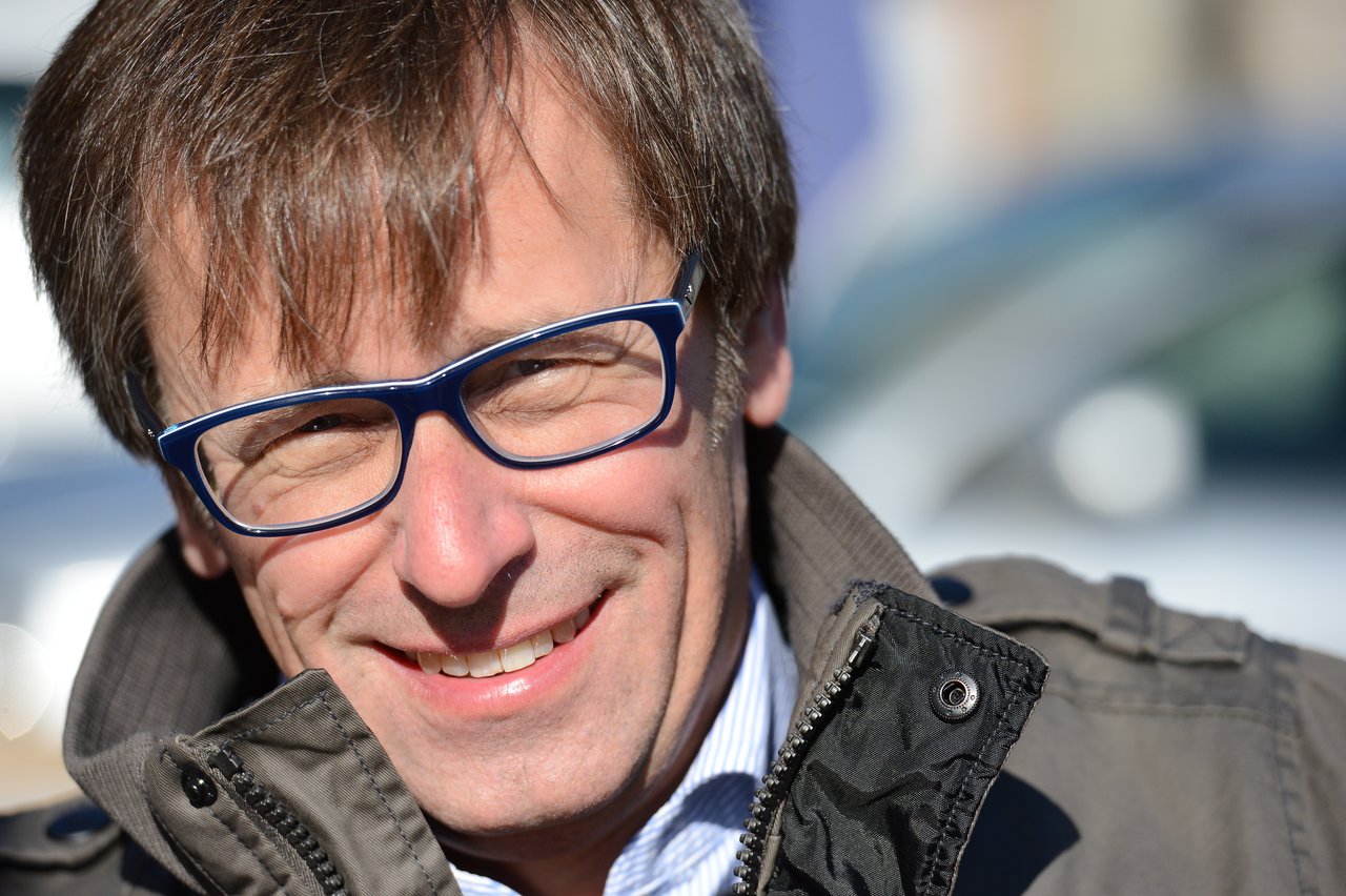 A man with blue glasses and a jacket smiles while looking at the camera in bright sunlight.