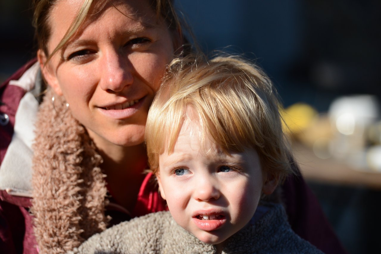 A woman and a young child sit closely together outdoors, both looking in different directions with neutral expressions.