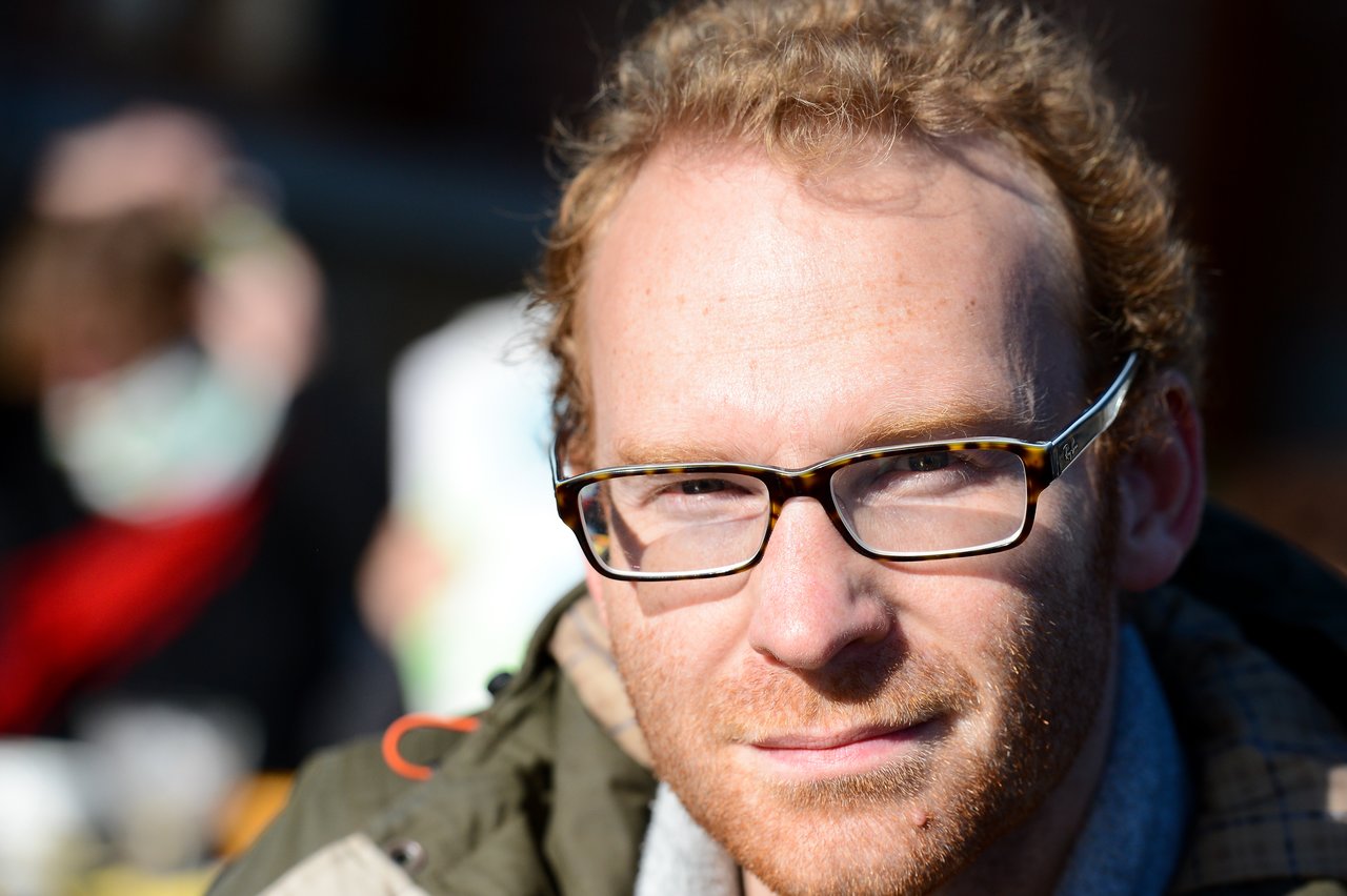 A man with glasses and curly hair looks directly at the camera, wearing a jacket in bright sunlight.