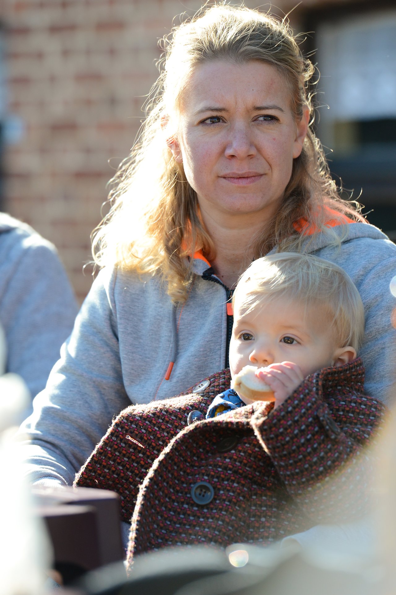 A woman in a gray hoodie holds a baby in a coat while the baby eats a piece of bread.
