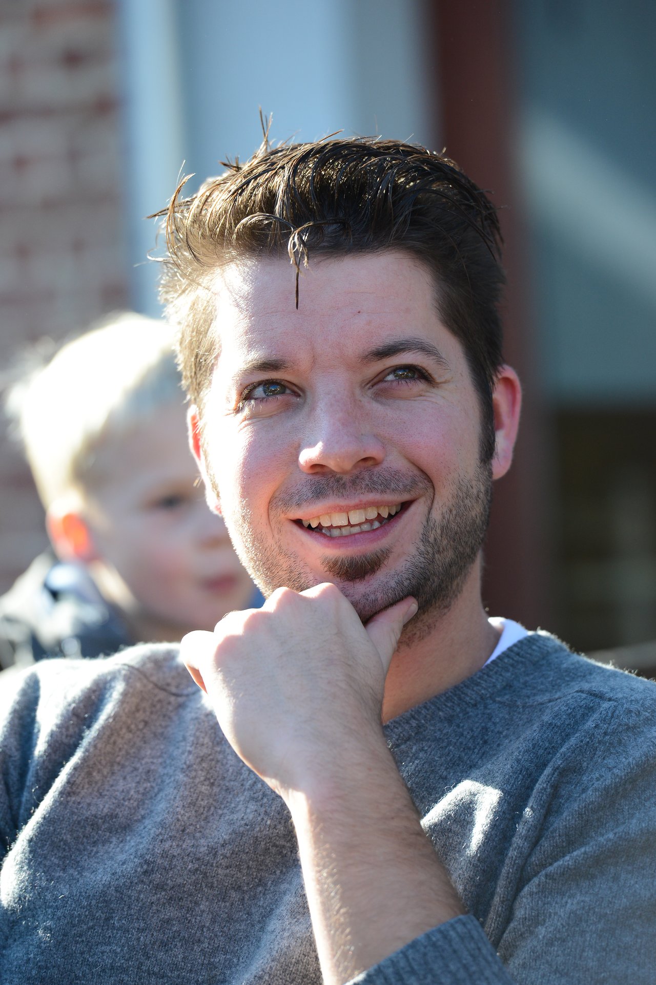 A man in a gray sweater smiles while resting his chin on his hand.