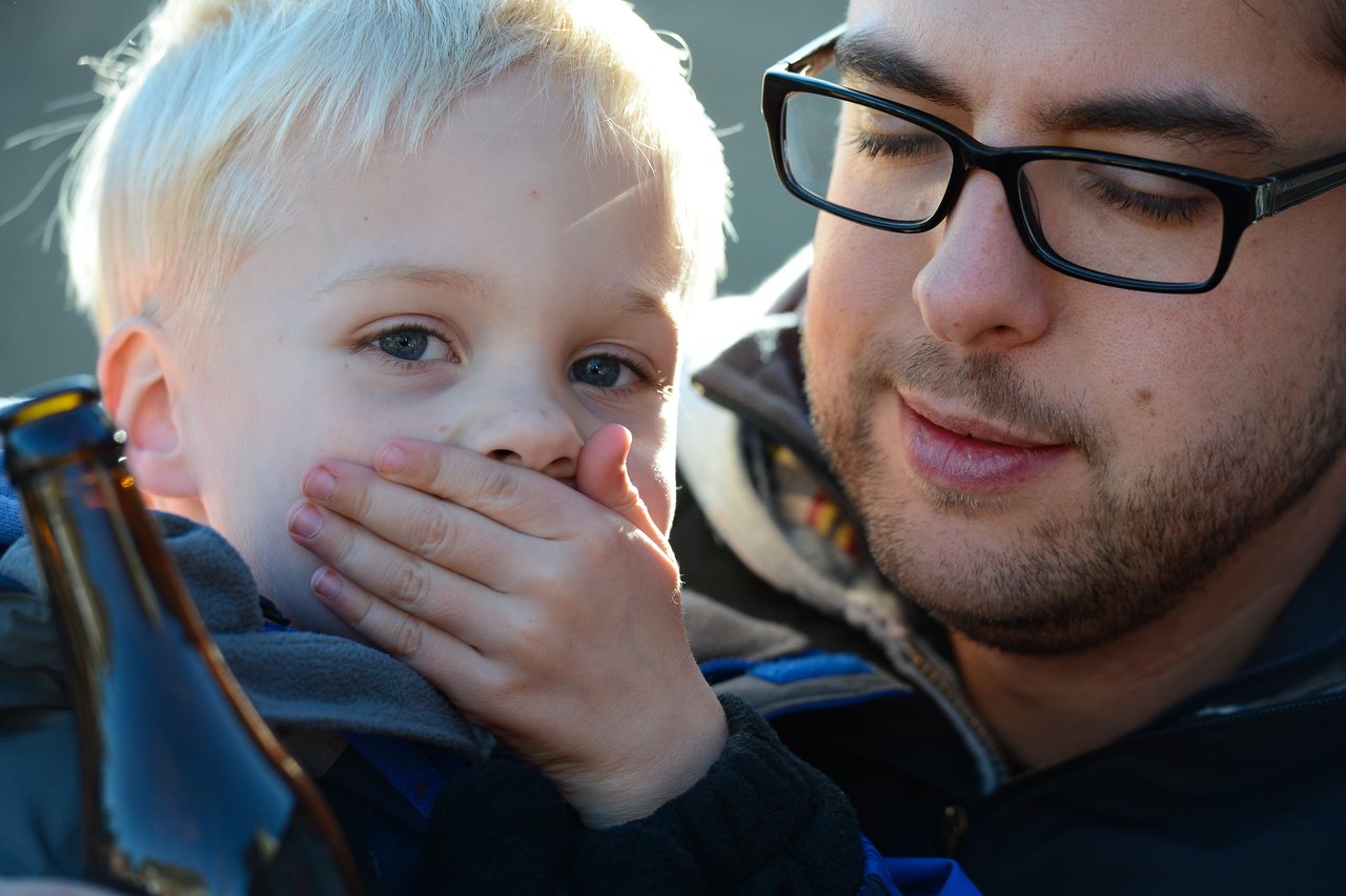 A young child covers their mouth with one hand while sitting close to an adult wearing glasses.