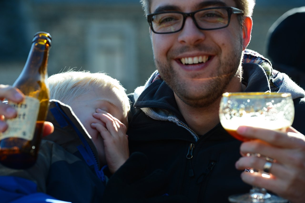 A smiling man holds a glass of beer while a child leans against him, covering their face with a hand.