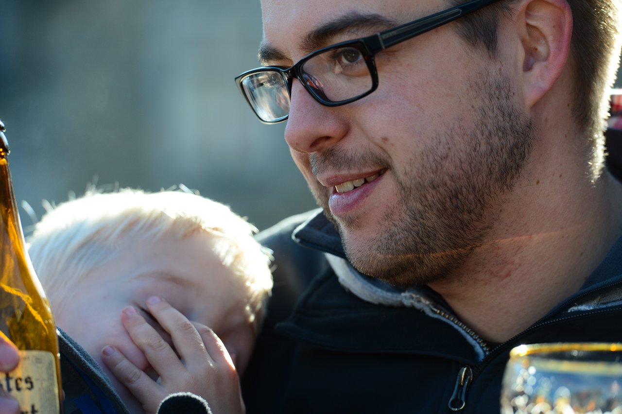 A man wearing glasses smiles while holding a young child who rests their head on his chest.
