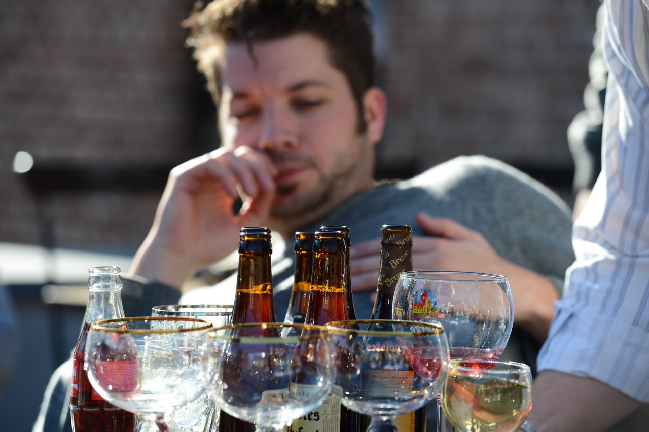 A man leans back while surrounded by bottles and glasses filled with drinks at an outdoor gathering.
