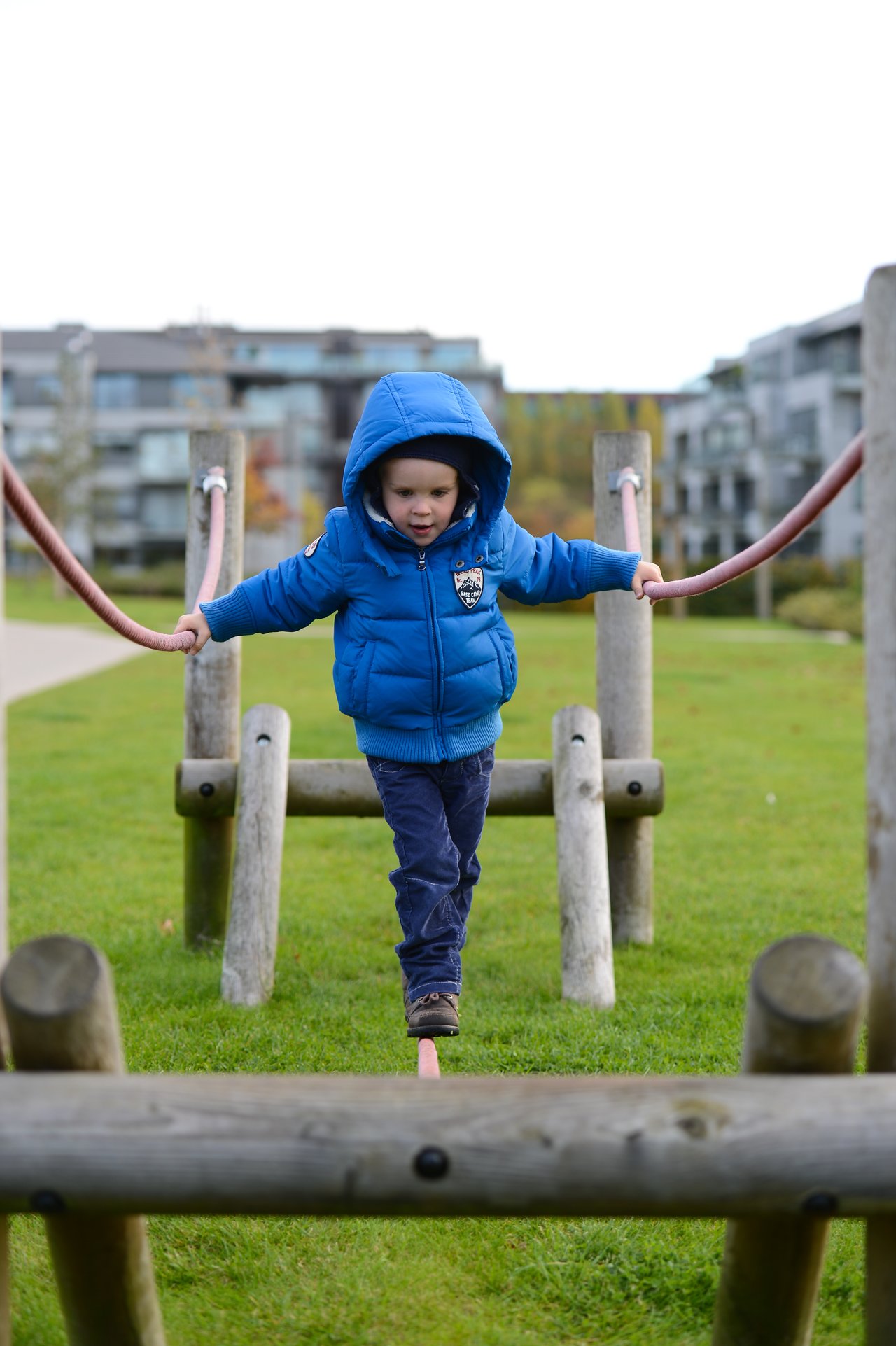 A young child in a blue jacket carefully walks on a balance beam, holding onto ropes for support.