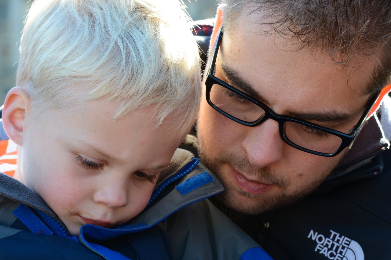 A man wearing glasses looks closely at a young child in a jacket, both appearing focused on something.