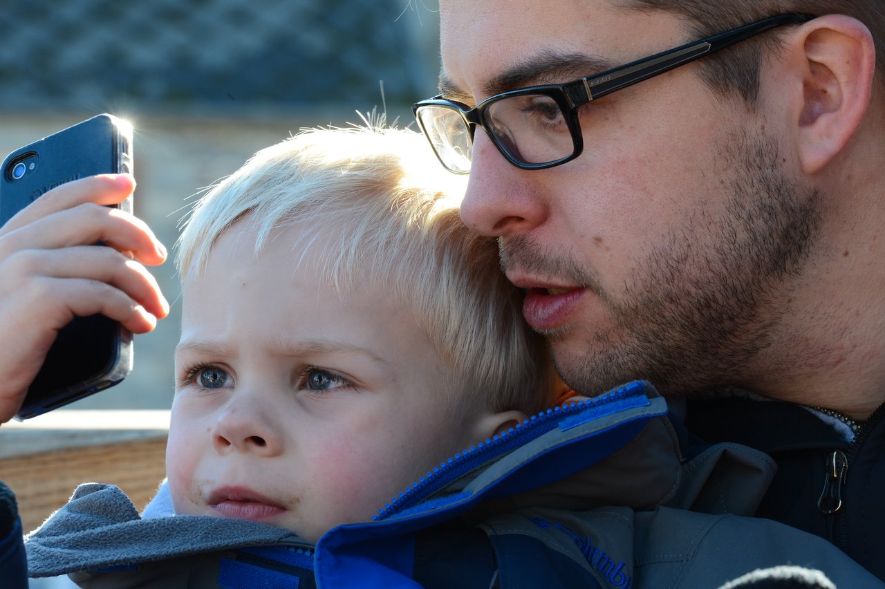 A young child holds a smartphone while an adult leans in closely, both looking at the screen intently.