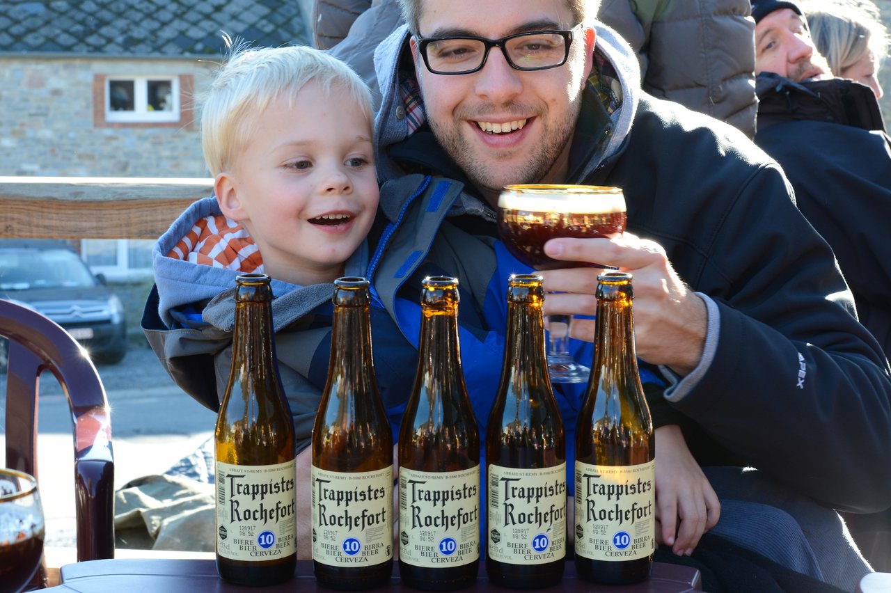 A man holding a glass of beer sits with a smiling child behind several bottles of Trappistes Rochefort beer.