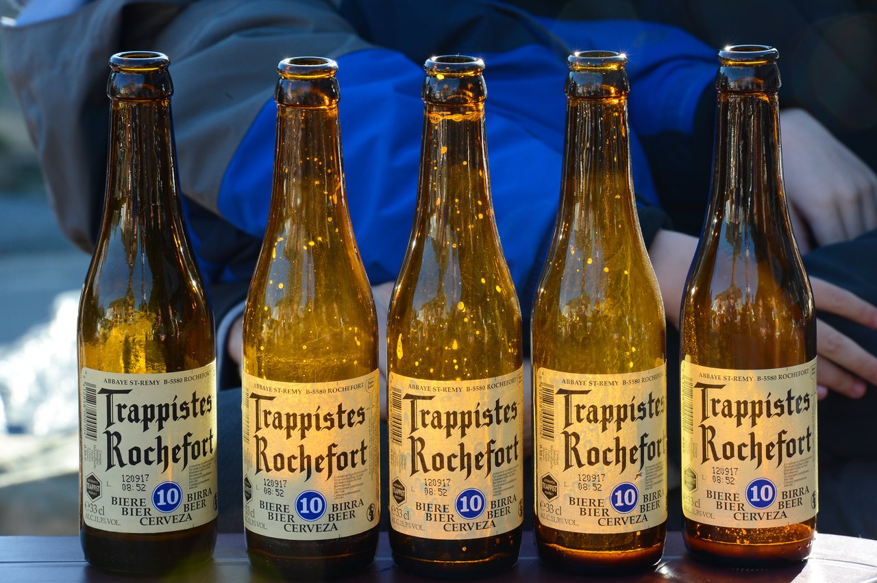 Five empty Trappistes Rochefort beer bottles are lined up on a table, with people sitting in the background.