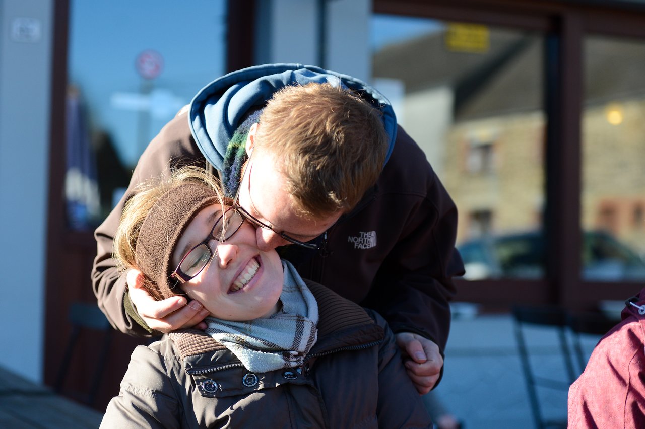 A man in a jacket leans in and kisses a smiling woman on the cheek outdoors.