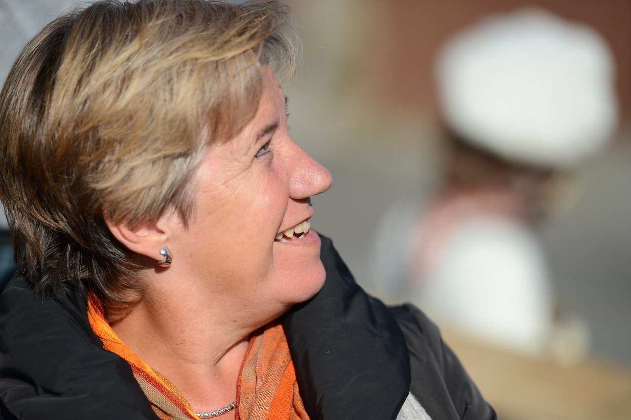 A woman with short hair and an orange scarf smiles while looking to the side.