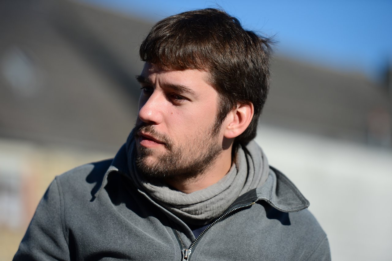A man with short brown hair and a beard looks to the side, wearing a gray jacket and scarf outdoors.