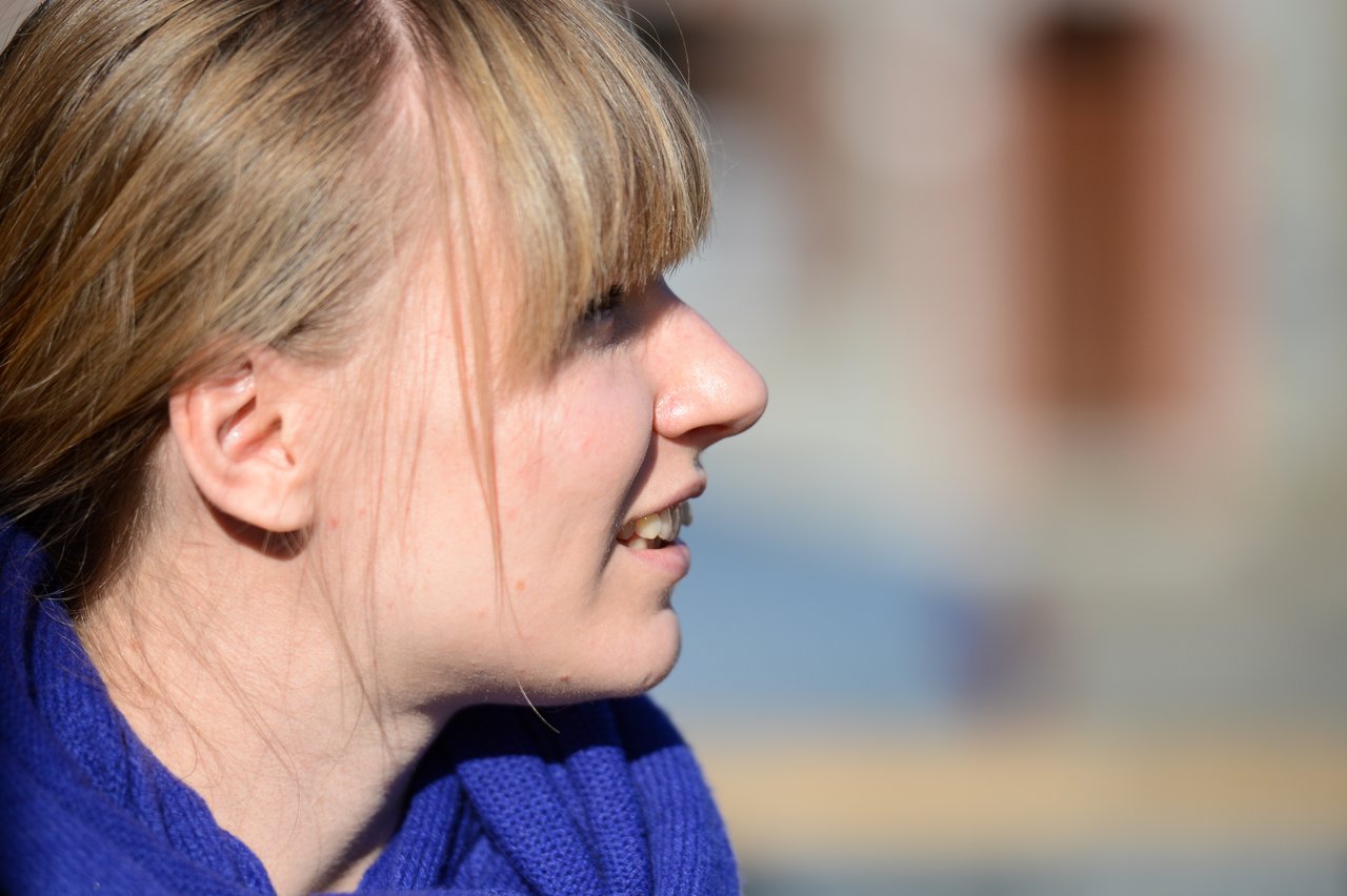 A woman with blonde hair and a blue scarf looks to the side and smiles in bright sunlight.